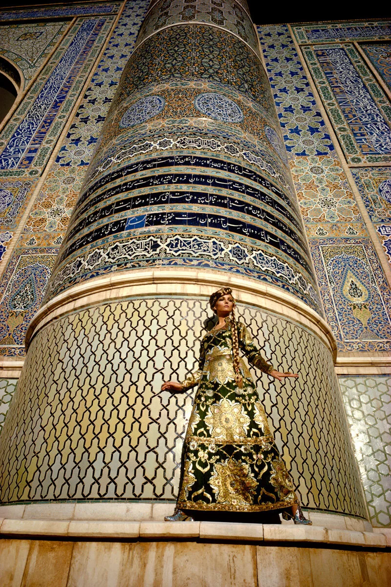 Model in an embroidered green dress standing against a tiled Islamic pillar.